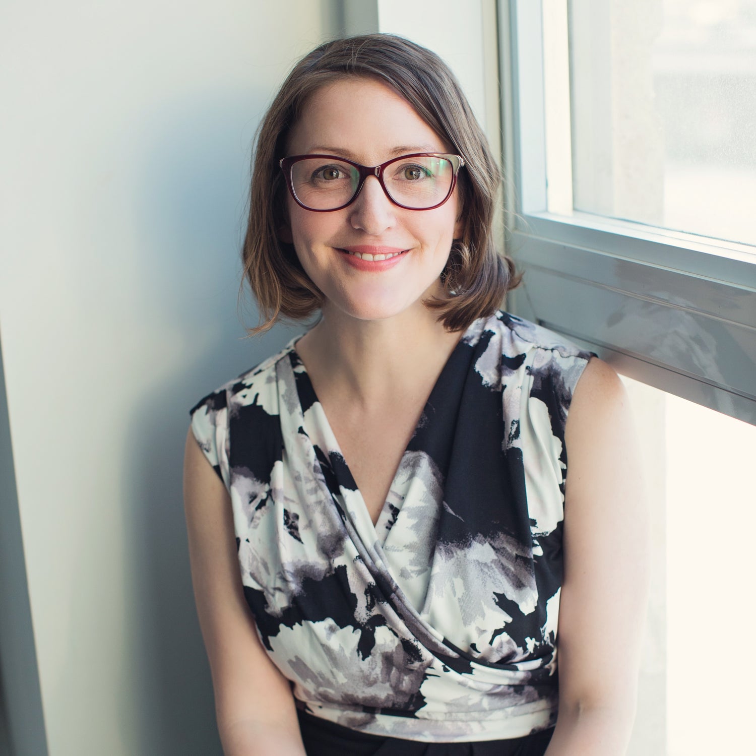 Portrait photo of woman wearing glasses and black-and-white floral dress in corner against bright window and wall